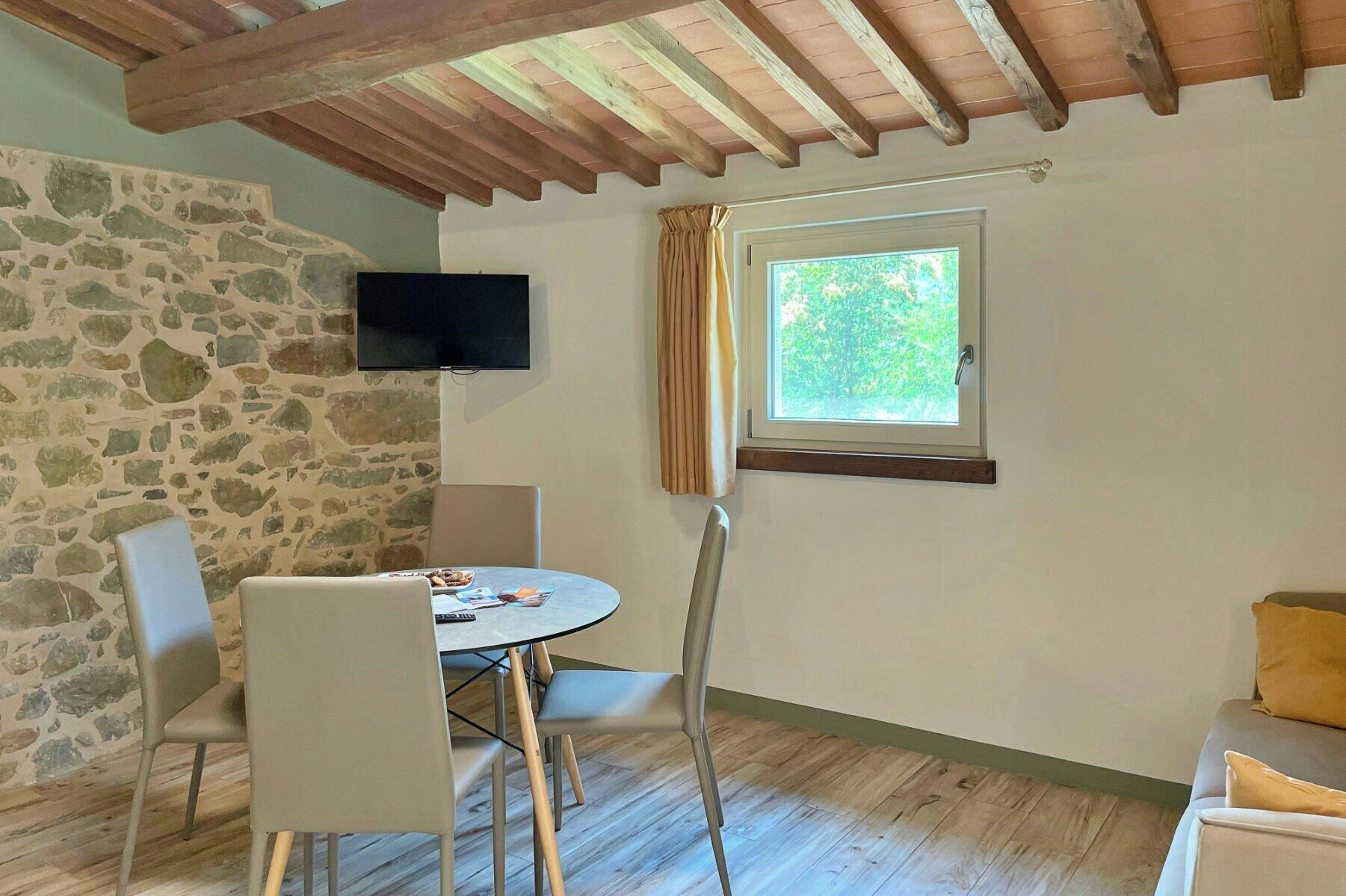 Ein Zimmer mit Steinwand, einem Tisch mit Stühlen, einem Fernsehgerät darüber, einem fenster mit Vorhängen und einer Holzbalkendecke.