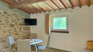 Habitació amb paret de pedra, televisió penjada sobre una taula amb cadires, finestra amb cortines i sostre de fusta.