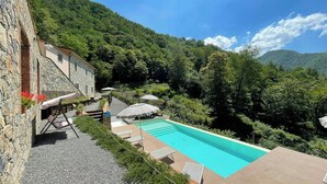 Edifici de pedra amb piscina, envoltat de vegetació abundant i turons.