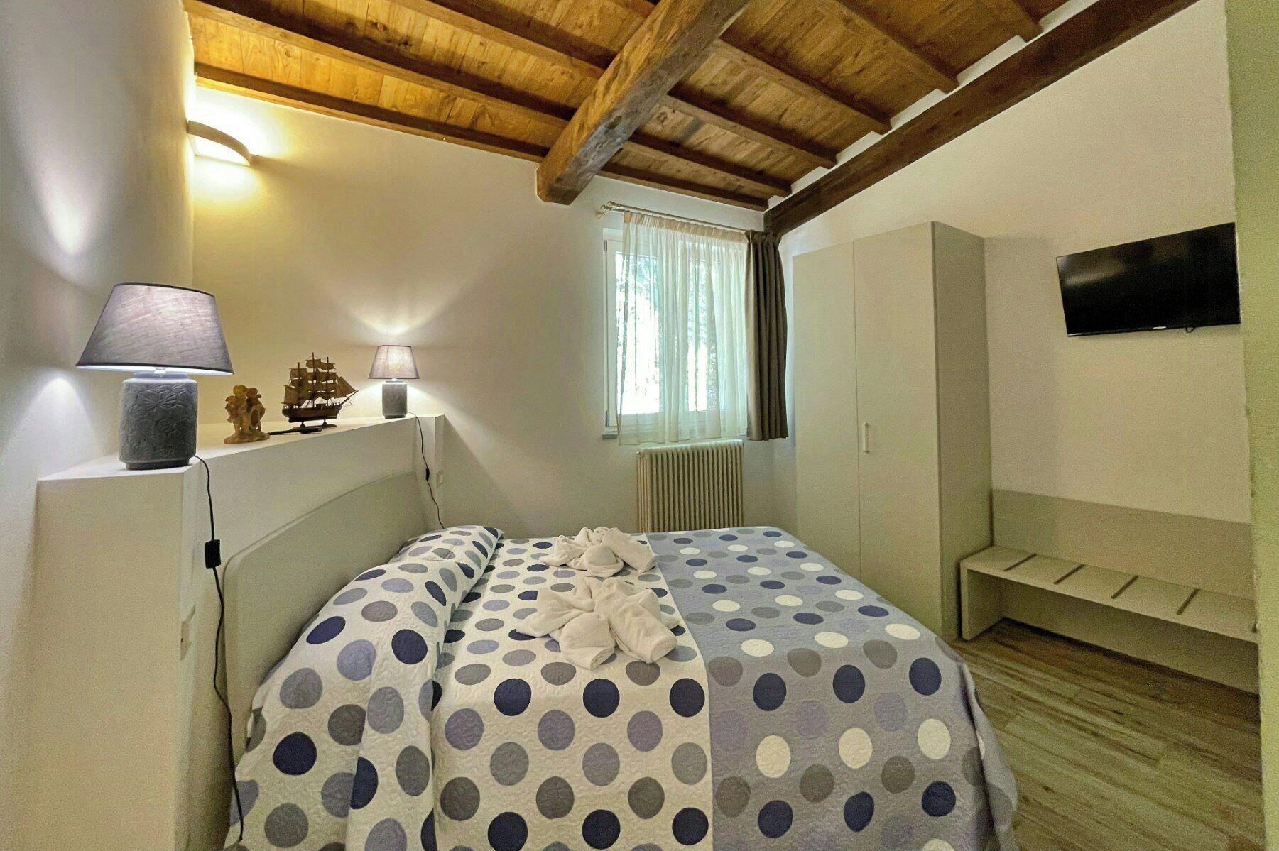 A bedroom with a bed, a nightstand, a lamp, a ship model, a window with curtains, and a TV on the wall.