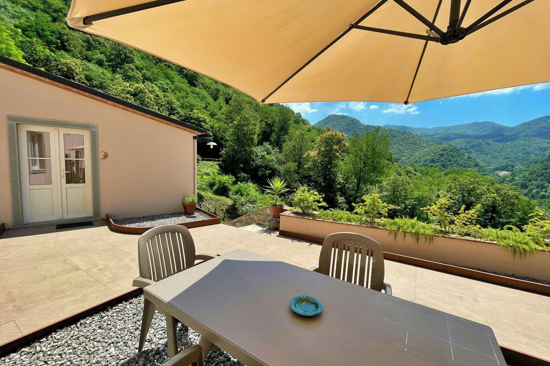 Un patio al aire libre con una mesa, sillas, un paraguas color beige y vistas a un paisaje montañoso.