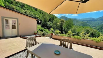 Un patio al aire libre con una mesa, sillas, un paraguas color beige y vistas a un paisaje montañoso.