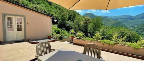 Un patio al aire libre con una mesa, sillas, un paraguas color beige y vistas a un paisaje montañoso.