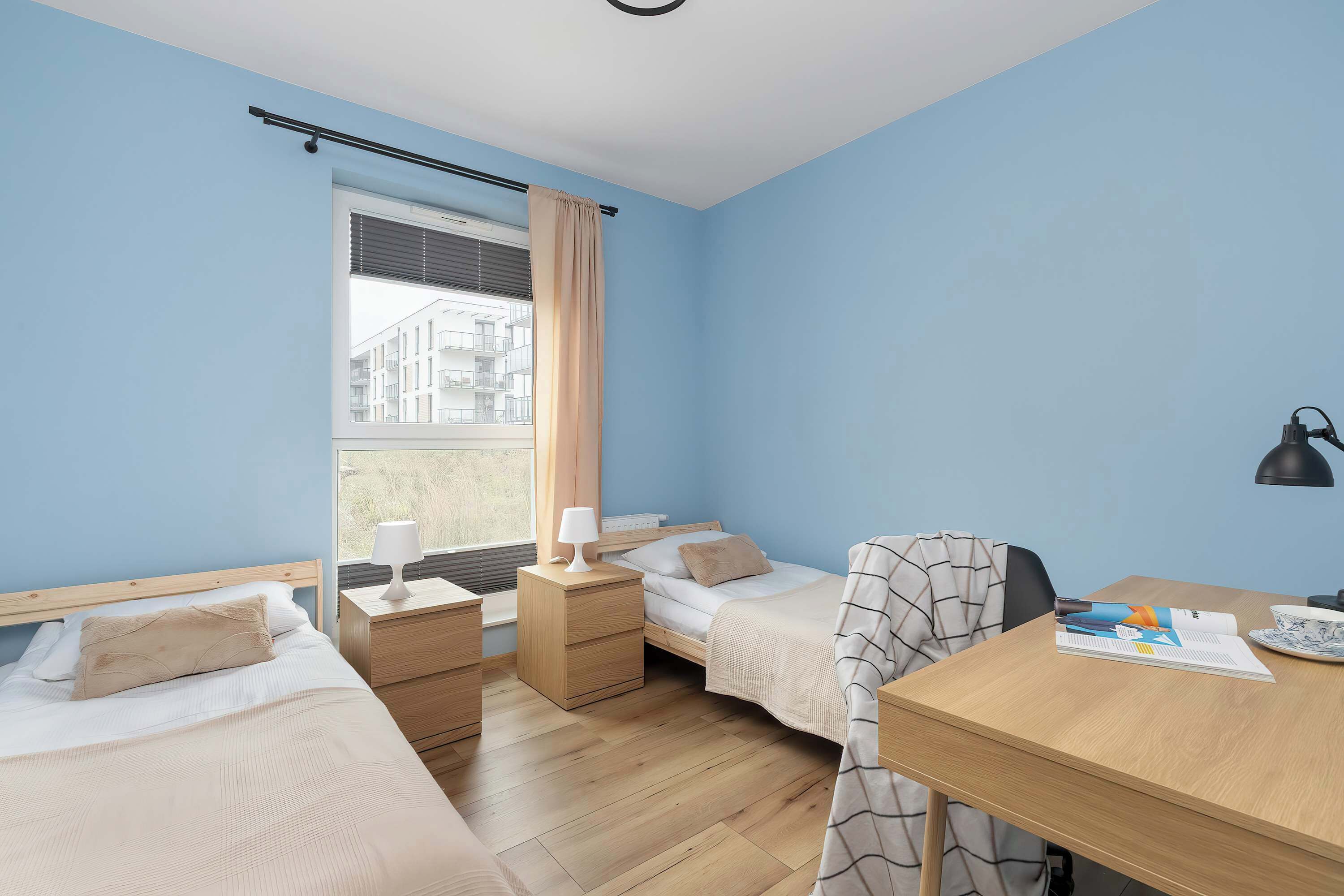 Ett hotellrum med två separata sängar, ett skrivbord i trä, en stol, en lampa och utsikt över byggnader genom fönstret.