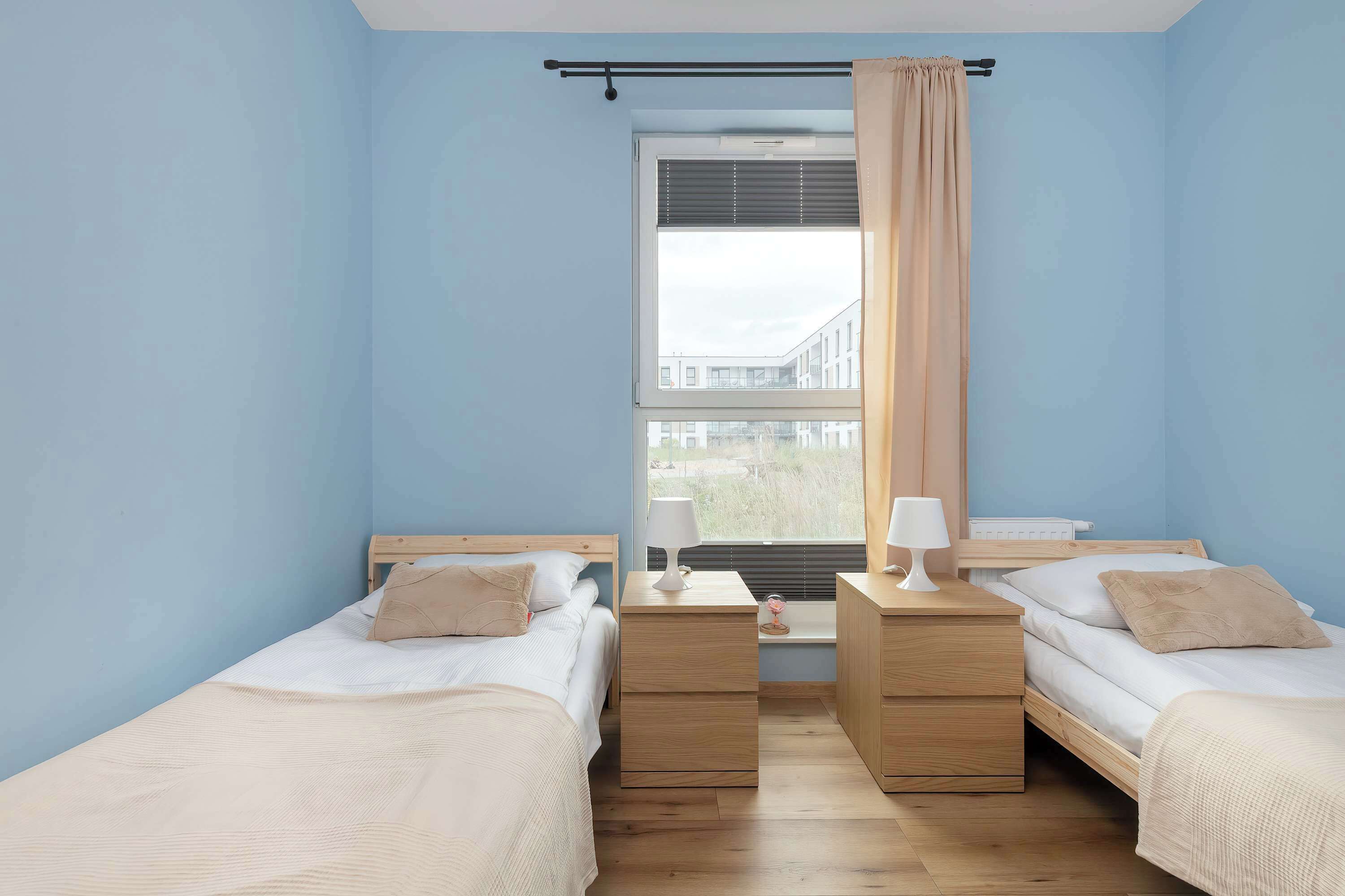 Un dormitorio con dos camas, una ventana con persianas, una mesita de noche con lámpara y vistas a edificios.