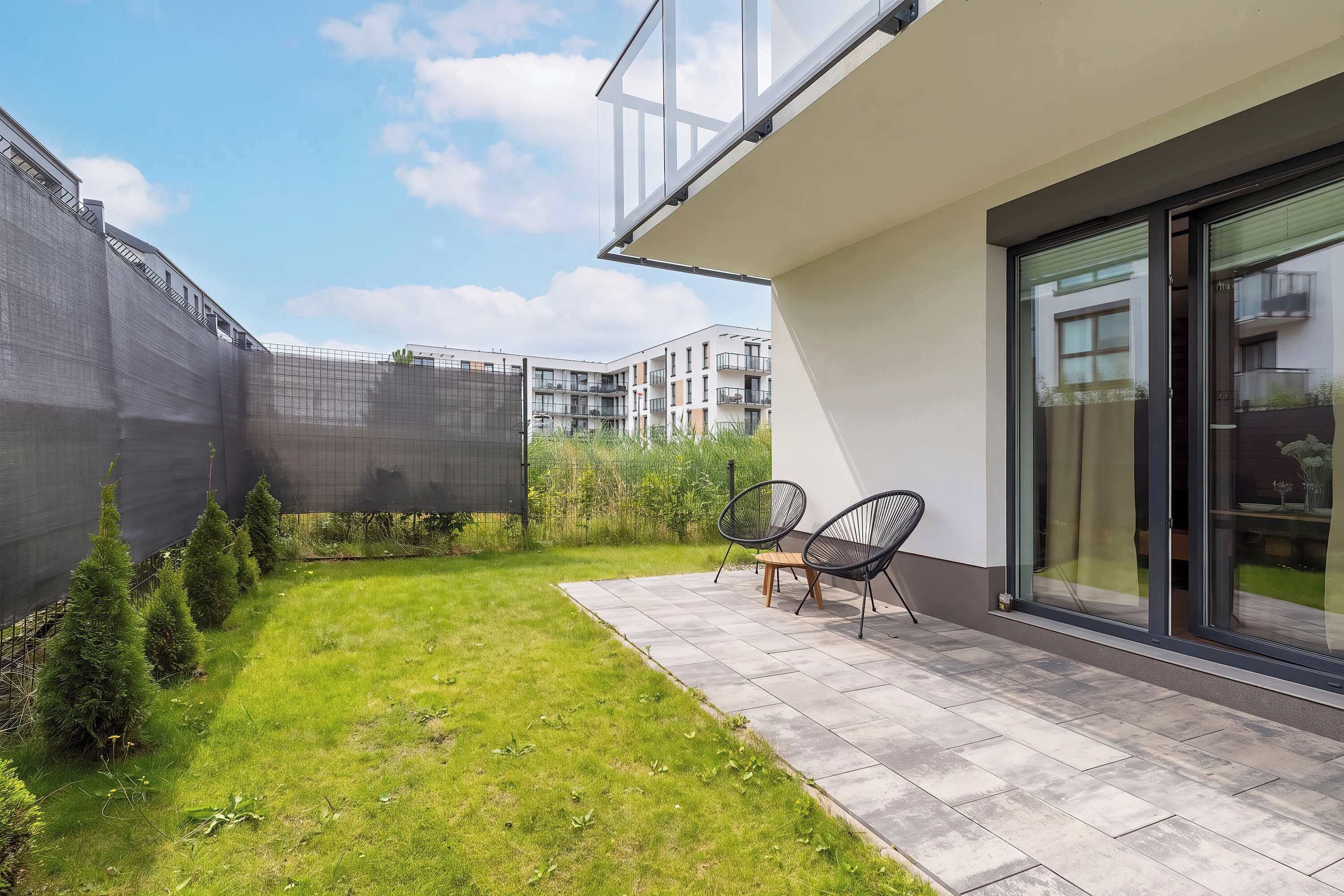 Un edificio residencial moderno con un patio empedrado, dos sillas exteriores y vistas a otros edificios de apartamentos.