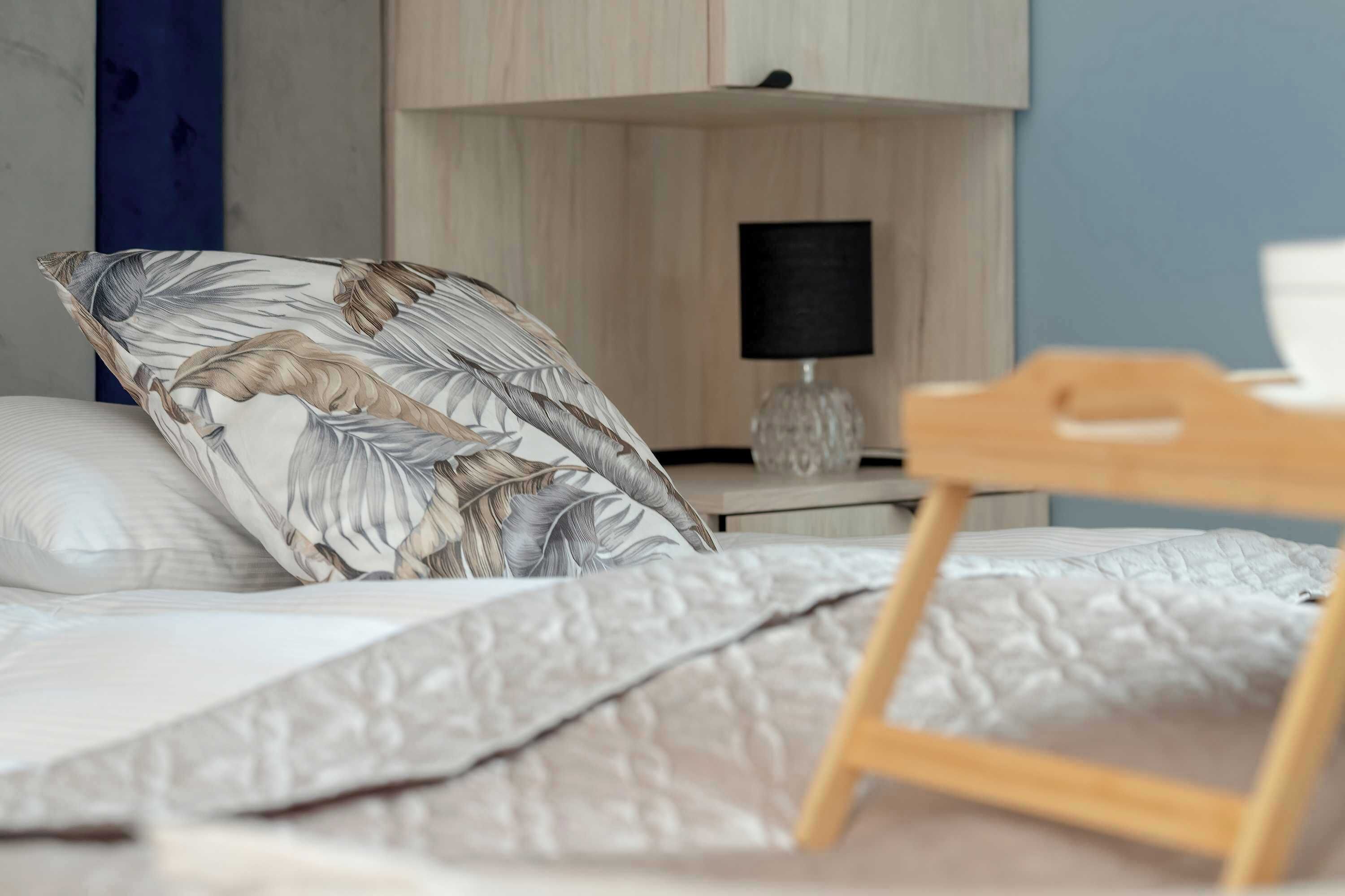Un letto rifatto con cura, un cuscino a fiori, un comodino con una lampada e un tavolino con una sveglia.