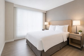 Ein Hotelzimmer mit einem großen Bett, zwei Nachttischen inklusive Lampen, einer kleinen Pflanze und einem Fenster mit Jalousien.