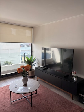A modern living room with a large flat-screen TV, a round table with a vase of flowers, and a black entertainment unit. - Charming 1-bedroom apartment in pleasant Concepción (Concepción)