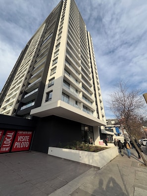 A high-rise building with multiple floors and balconies, a signboard with contact information, and a paved walkway with people walking. - Charming 1-bedroom apartment in pleasant Concepción (Concepción)