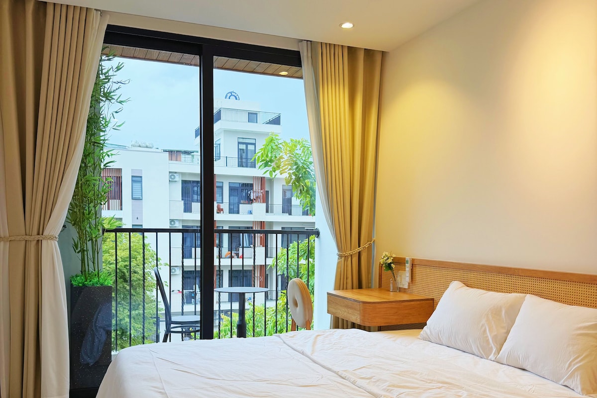 A hotel room with a large bed, a wooden headboard, a nightstand, a potted plant, and a view of a building through the window.