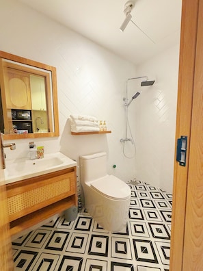 A modern bathroom with a white toilet, a wooden vanity, a mirror, and a shower. - La Van Residence (Da Nang)