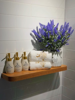 A bathroom shelf with La Van Residence toiletries, a vase of purple flowers, and neatly folded towels. - La Van Residence (Da Nang)