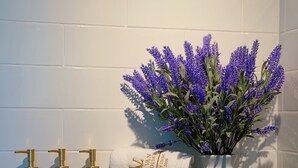 A bathroom shelf with toiletries, a rolled towel, and a vase with purple flowers. - La Van Residence (Da Nang)