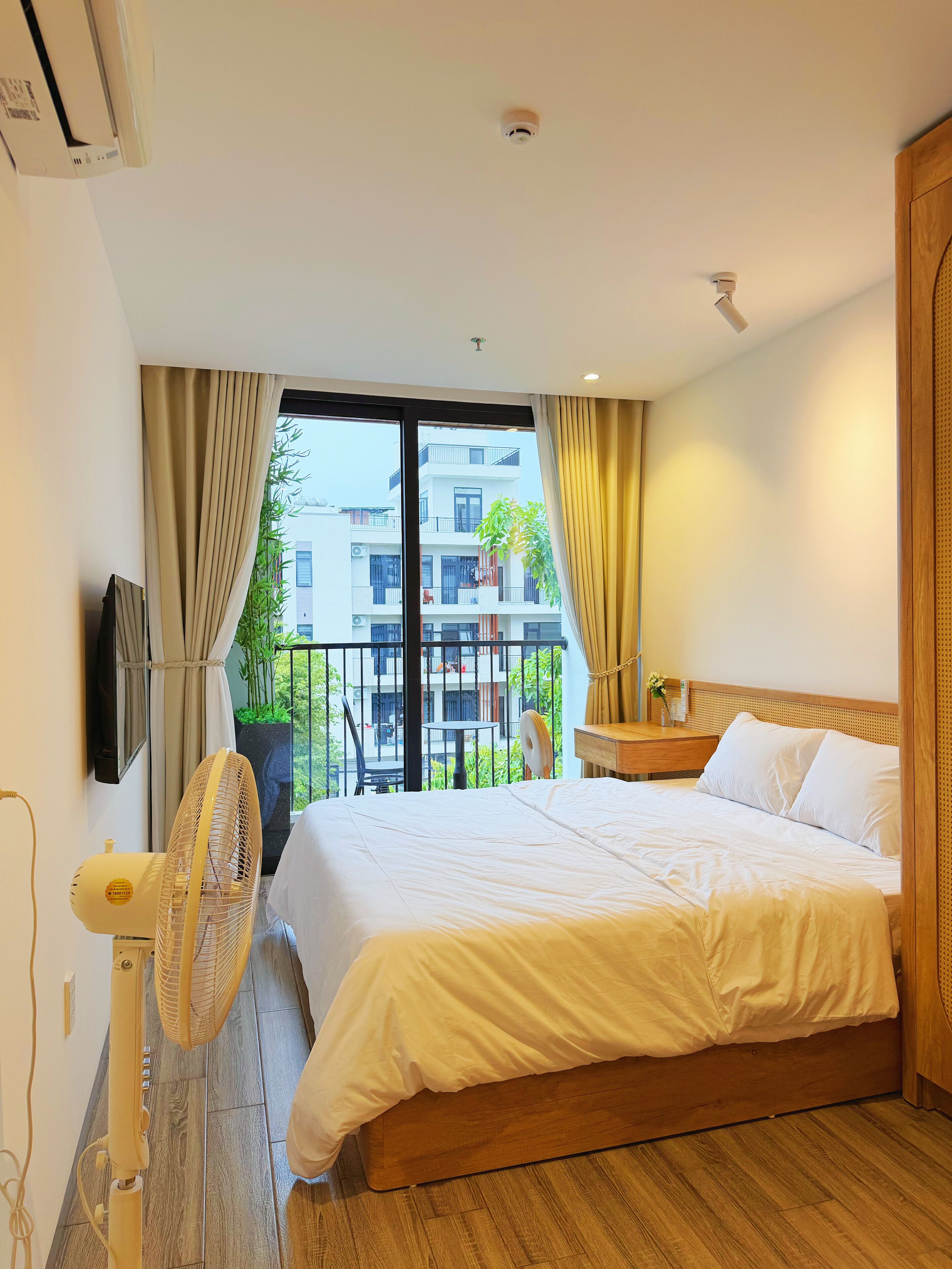 Habitación de hotel con cama, mesita de noche, ventilador, televisor y vistas al balcón exterior.