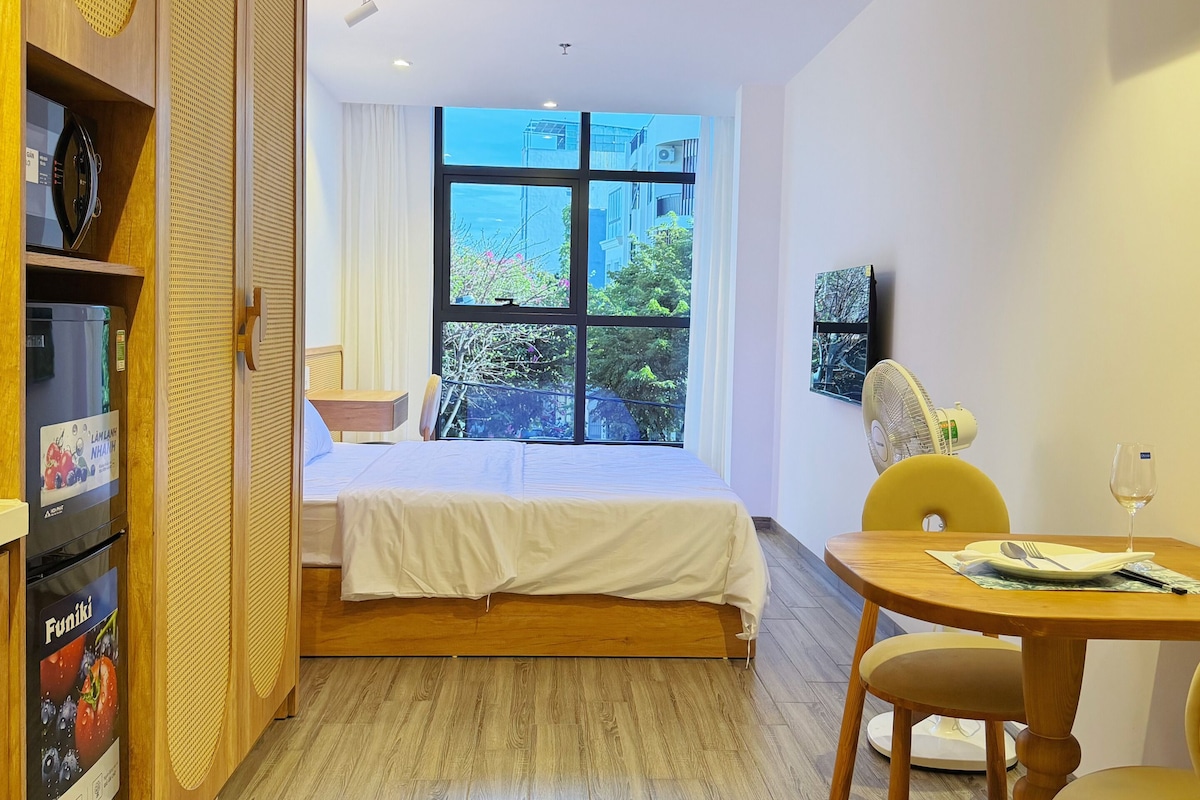 A hotel room with a bed, a dining table with chairs, a kitchenette, and a large window offering a view of buildings and trees.