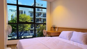 Habitación de hotel con un ventanal grande, una cama, una mesita de noche con un florero y un ventilador de pie.