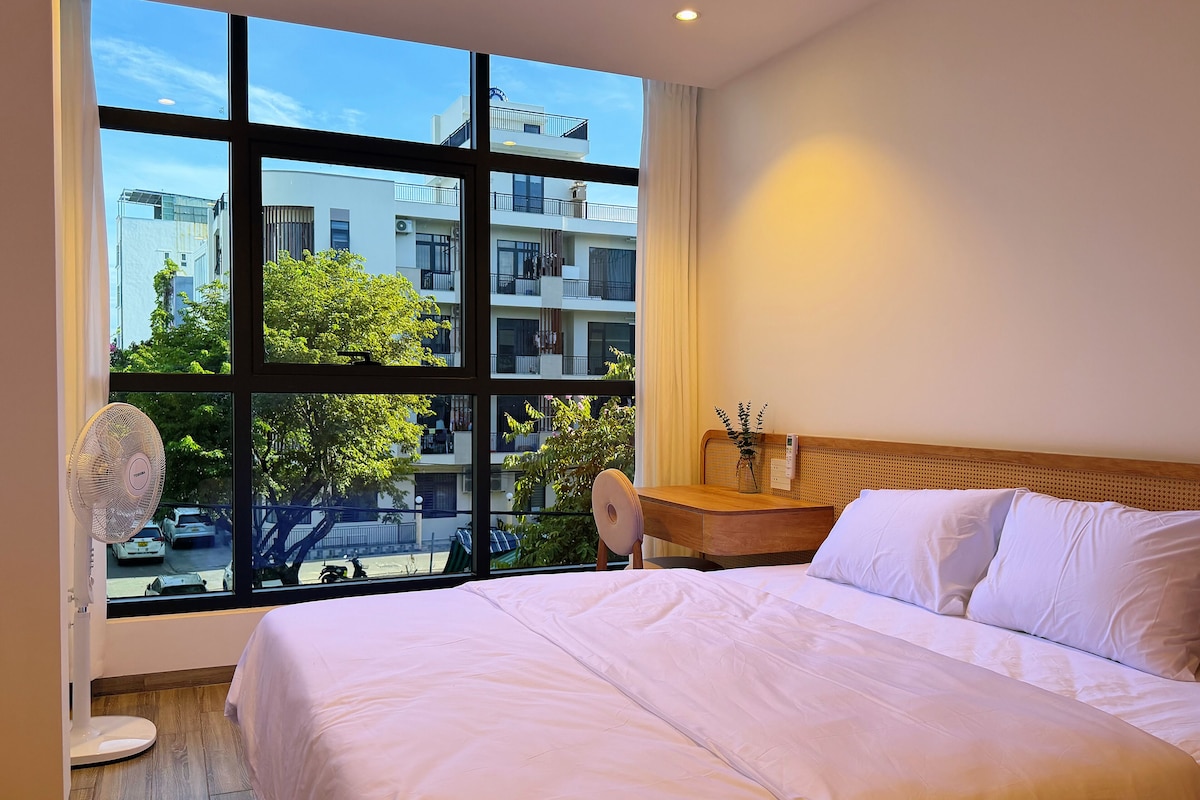 A hotel room with a large window, a bed, a bedside table with a vase, and a standing fan.