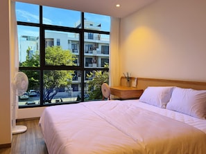 A hotel room with a large window, a bed, a bedside table with a vase, and a standing fan. - La Van Residence (Da Nang)