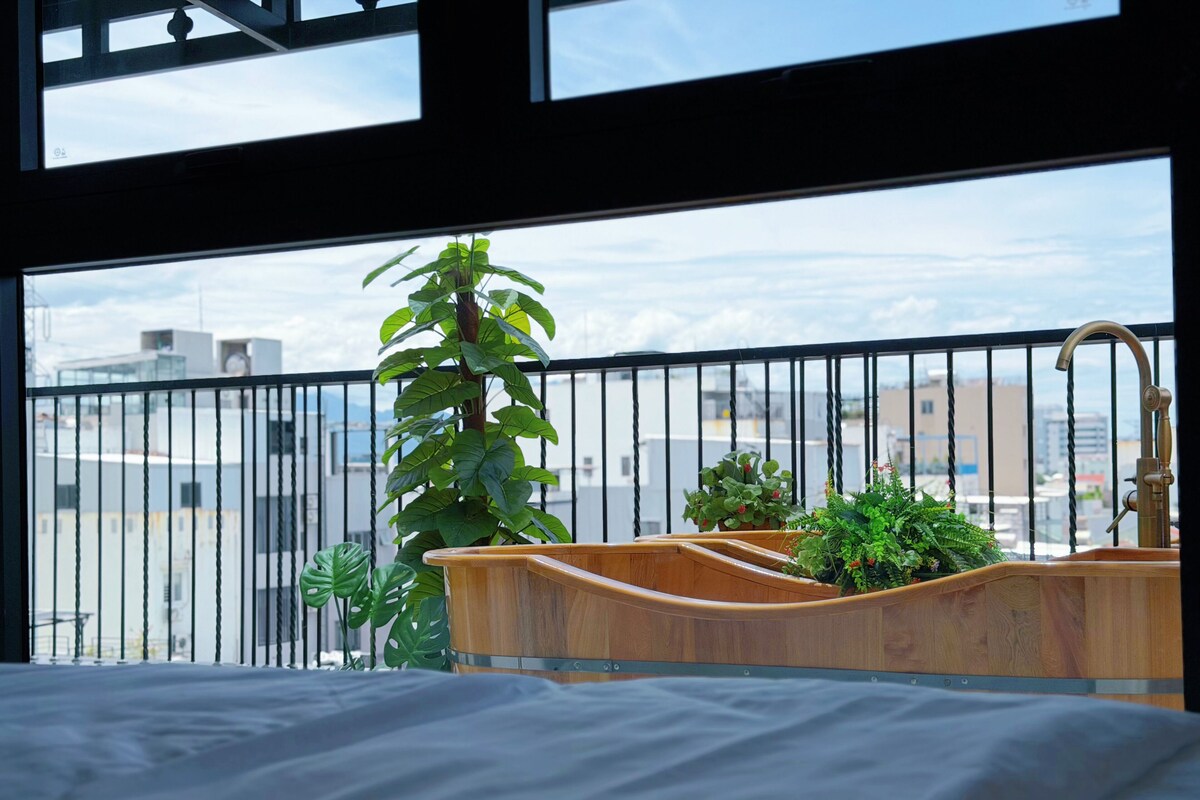 A bedroom with a wooden bed, a wooden sink, and a view of the city through a balcony with a black railing.
