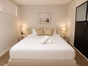 Una cama bien hecha con una toalla enrollada, dos lámparas de noche y un cuadro en la pared.