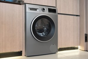 A modern, stainless steel front-loading washing machine in a kitchen with wooden cabinets and a tiled floor. - StayShort - Cozy Studio in Jumeirah Garden City with Balcony Sleeps 2 (Jumeirah Garden City)