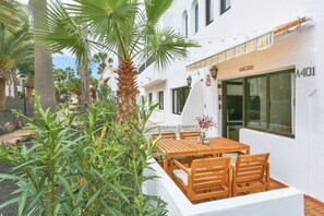 A white building with a wooden table and chairs outside, surrounded by plants. - Brand New Apt - Oasis Duna pools (Corralejo)