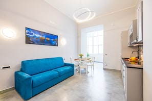 Ein moderner Wohnraum mit einem blauen Sofa, einem Esstisch mit Stühlen, einer Küchenzeile mit Schränken und einem großen Fenster, das für natürliches Licht sorgt.