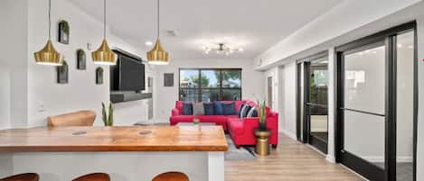 Una cocina moderna con una isla central, taburetes de bar, iluminación colgante y un sofá rojo en la sala de estar.