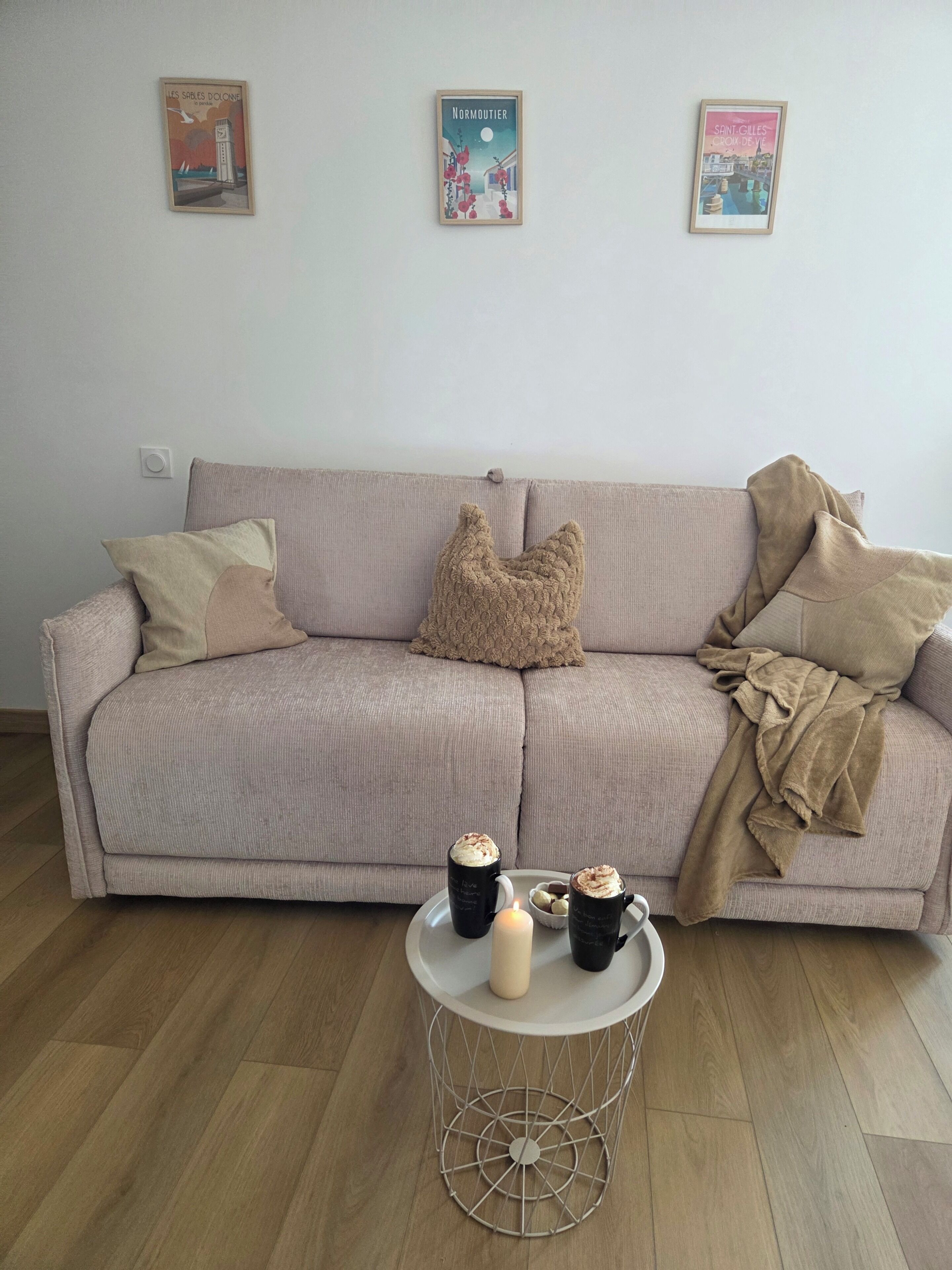 A living room with a beige sofa, a round coffee table with two mugs and a candle, and three framed pictures on the wall.