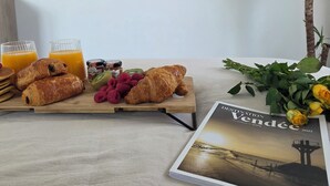 A breakfast setup with croissants, fruits, and juice on a wooden board, a magazine, and a TV displaying a beach scene. - Cozy beachfront studio (Brem-sur-Mer)