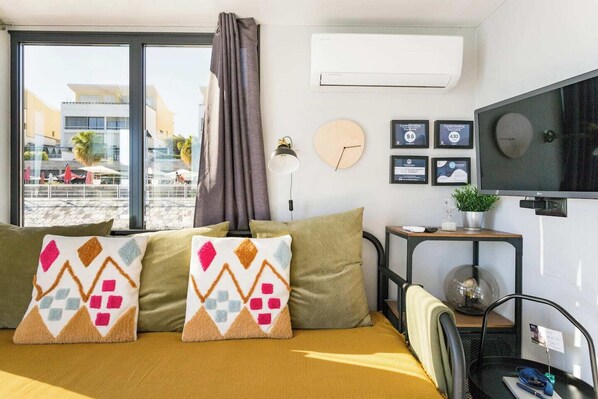A modern hotel room with a large window, a flat-screen TV, a small table with a plant, and a bed with patterned pillows.