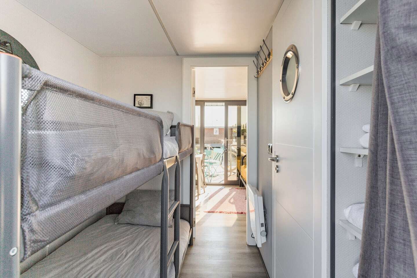 A narrow hallway with bunk beds on the left, a door leading to a balcony, and a bathroom with a towel rack and a round mirror on the right.