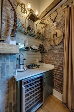 A kitchen with a brick wall, wooden shelves, a clock, and a whiskey barrel sign. - The Full Proof Penthouse in Downtown Bardstown - Walking to Restaurants & Shops! (Bardstown)