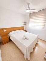 Un dormitorio con cama, mesita de noche, lámpara de pie, ventilador de techo y ventana con persianas.