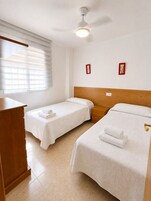 Habitación de hotel con dos camas individuales, cabecera de madera, mesita de noche, escritorio y ventilador de techo.