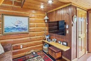 Ein Holzblockhaus-Innenraum mit einem angebrachten Fernseher, einem gerahmten Gemälde und einem Holzboden mit verschiedenen Gegenständen.