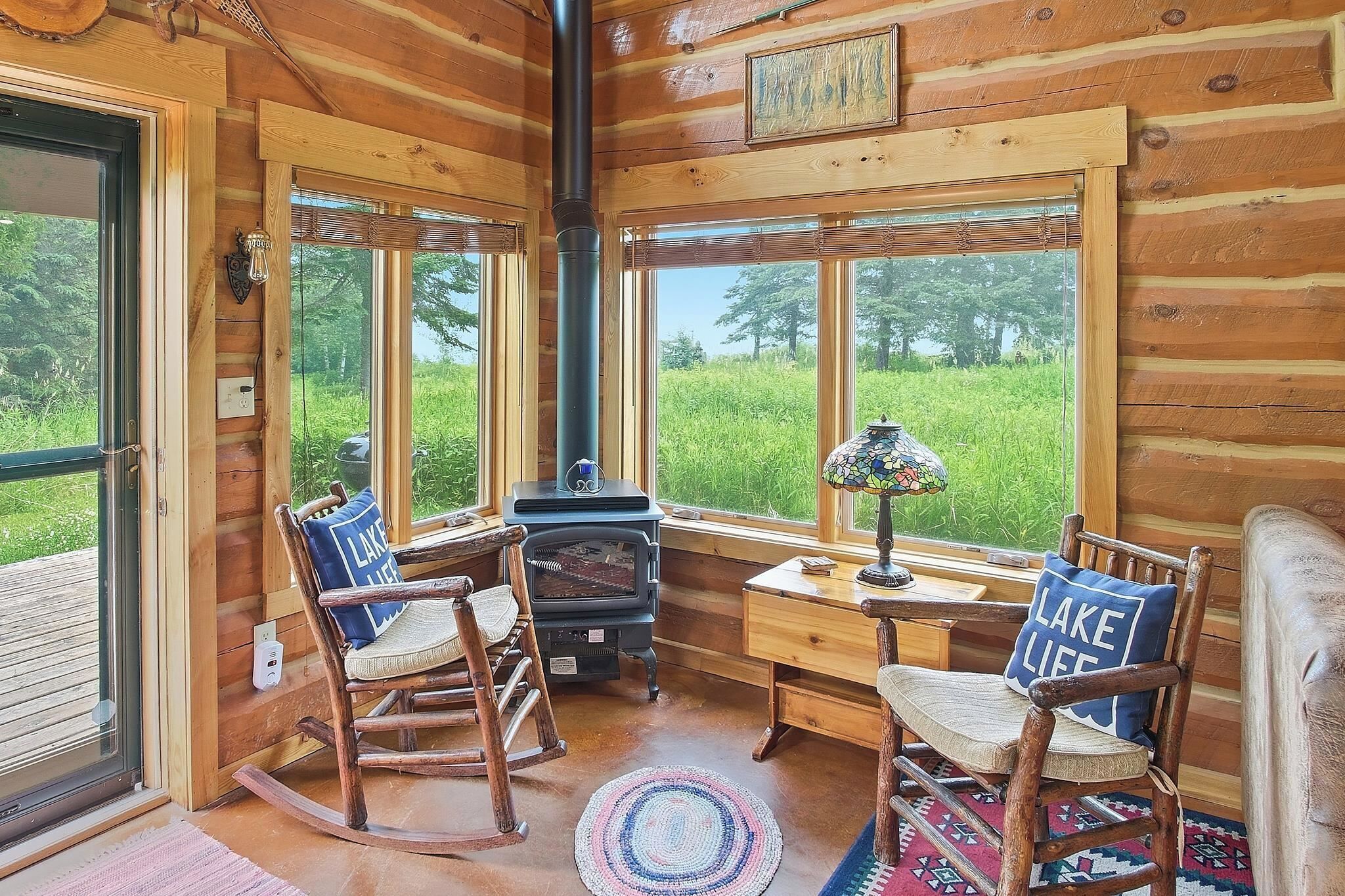 A cozy cabin interior with a wood stove, two rocking chairs, and a view of a green field.