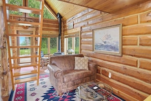 Ein gemütliches Blockhauss-Interieur mit einem Ledersofa, einem gemusterten Teppich und einem großen Fenster mit Blick ins Freie.