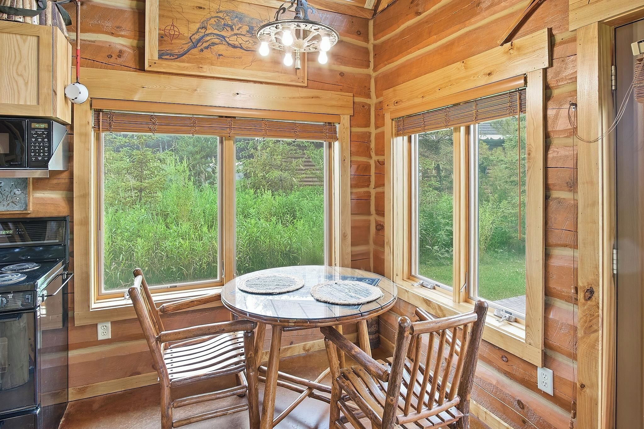 A wooden cabin interior with a round table and chairs, a stove, and large windows with a view of greenery.