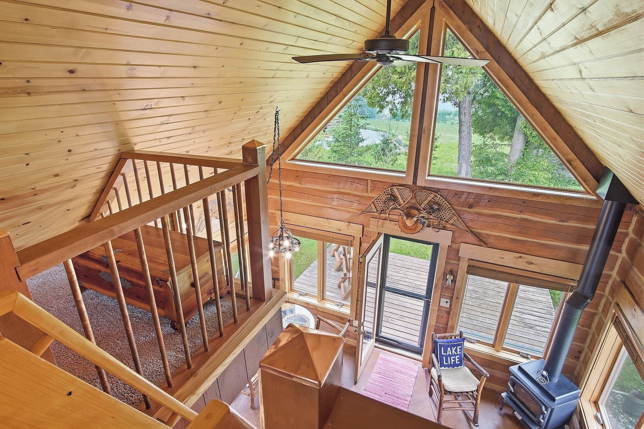 A wooden cabin interior with a loft bedroom, a fireplace, and a view of the outdoors.