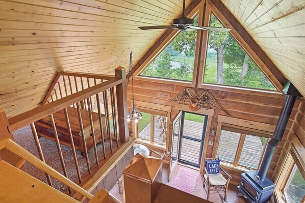 A wooden cabin interior with a loft bedroom, a fireplace, and a view of the outdoors.