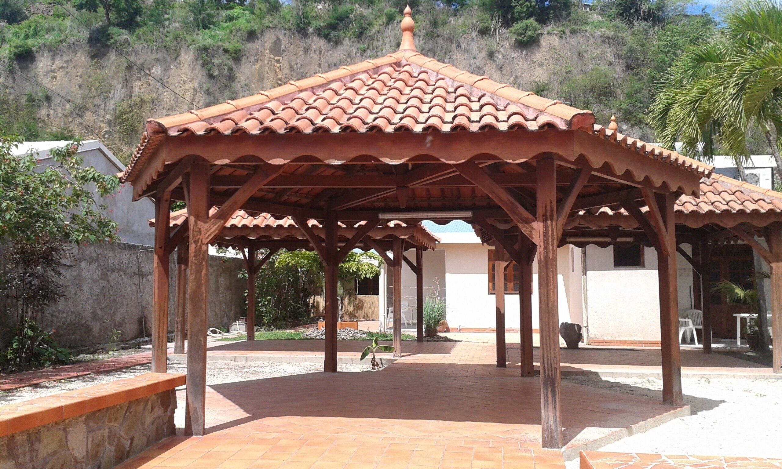 A gazebo with a tiled roof and wooden structure, surrounded by a paved area and greenery.