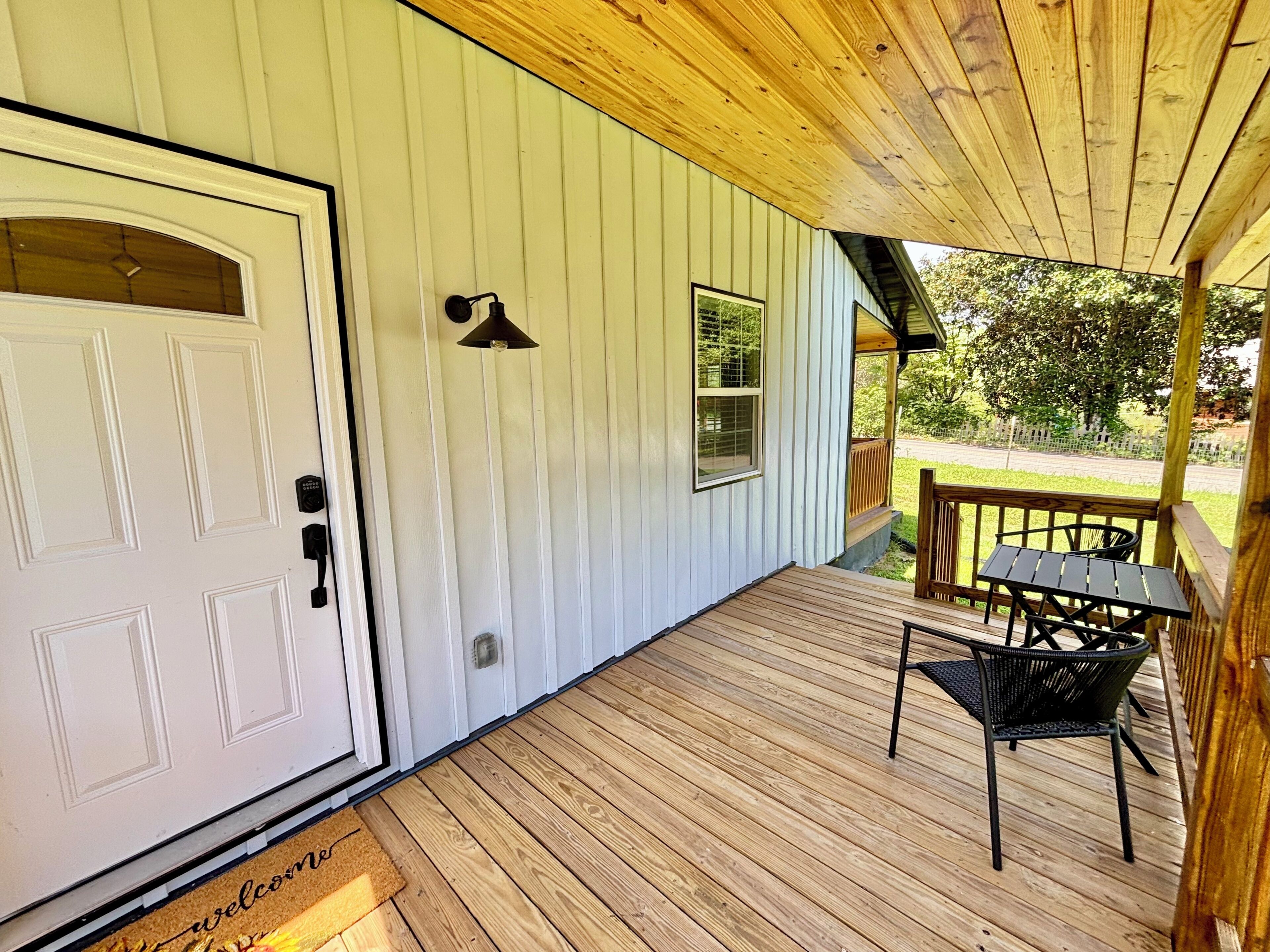 A wooden deck with a door, a welcome mat, a table with chairs, and a picture on the wall.