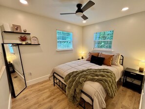A bedroom with a bed, a bedside table, a nightstand, a mirror, a ceiling fan, and two windows with blinds.