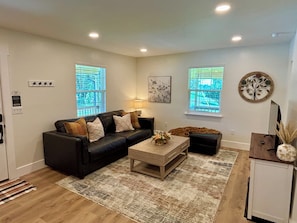 A living room with a black sofa, a wooden coffee table, and a rug. There are two windows with blinds, a wall-mounted lamp, and a decorative wall piece.