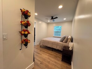 Une chambre à coucher avec un lit, un ventilateur de plafond, une fenêtre avec des stores et un arrangement floral fixé au mur.