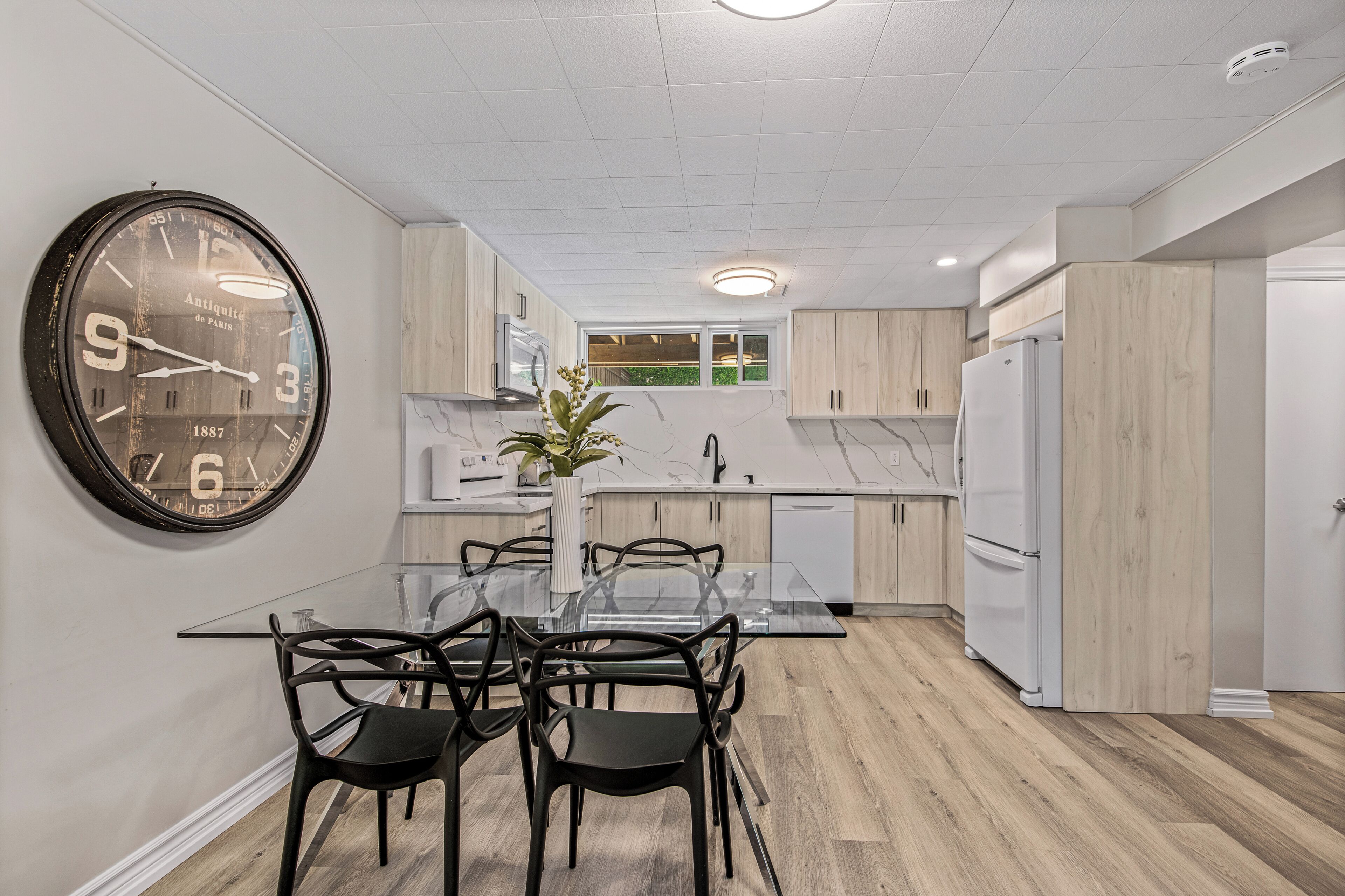 Una cocina moderna con un reloj de pared grande, armarios de madera, encimera de mármol y una zona de comedor con mesa y sillas de vidrio.