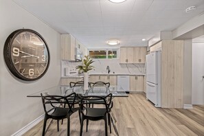 A modern kitchen with a large wall clock, wooden cabinets, a marble countertop, and a dining area with a glass table and chairs. - Beautifully Remodeled 2 Bedroom Basement Apartment (Toronto)