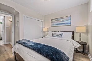A bedroom with a large bed, two bedside lamps, a painting on the wall, and a door leading to another room. - Beautifully Remodeled 2 Bedroom Basement Apartment (Toronto)